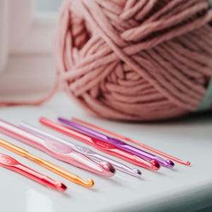 May include: A collection of colorful crochet hooks in shades of red, orange, pink, and purple, arranged in a row. A large ball of pink yarn is in the background, suggesting a crafting theme. The hooks are made of metal.