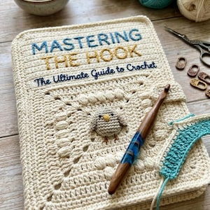 May include: A cream-coloured crocheted book cover with the text "MASTERING THE HOOK" and "The Ultimate Guide to Crochet". A wooden crochet hook and a small crocheted bird are on the cover. Yarn balls and a measuring tape are in the background.
