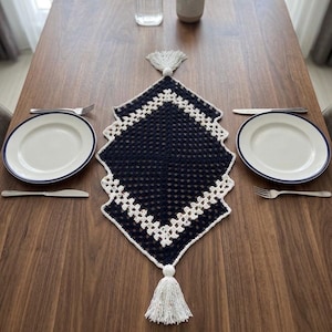 May include: A navy blue and white crocheted table runner with a diamond shape and tassel accents. The runner is placed on a wooden table, set with white plates, silverware, and a water pitcher.