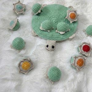Op de afbeelding: Een handgemaakt gehaakt schildpad speelgoed in groen, beige en oranje. De schildpad heeft een rond lichaam, een kop en verschillende kleine schildpadvormige stukken. Het speelgoed is gemaakt van zacht garen en is waarschijnlijk bedoeld voor kinderen.