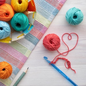 May include: A collection of colorful yarn balls in shades of orange, yellow, teal, and blue, arranged with a blue crochet hook and a pencil on a light wood surface. A heart shape is formed with red yarn.