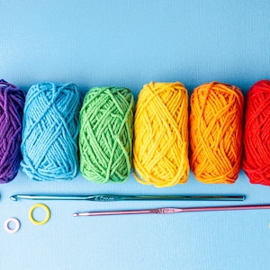 May include: A vibrant display of colorful yarn skeins in purple, blue, green, yellow, orange, and red. Two crochet hooks and stitch markers are also visible. The image is set against a light blue background, perfect for craft projects.