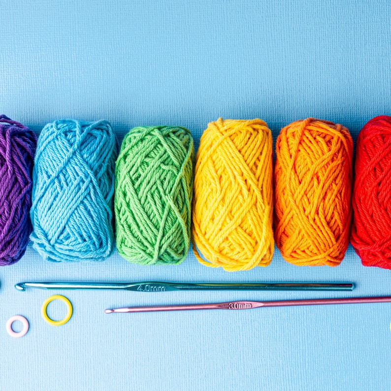 May include: A vibrant display of colorful yarn skeins in purple, blue, green, yellow, orange, and red. Two crochet hooks and stitch markers are also visible. The image is set against a light blue background, perfect for craft projects.
