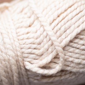 May include: Close-up of a ball of off-white yarn. The yarn is thick and textured, with visible strands tightly wound together. The color is a soft, creamy white, ideal for knitting or crocheting projects.