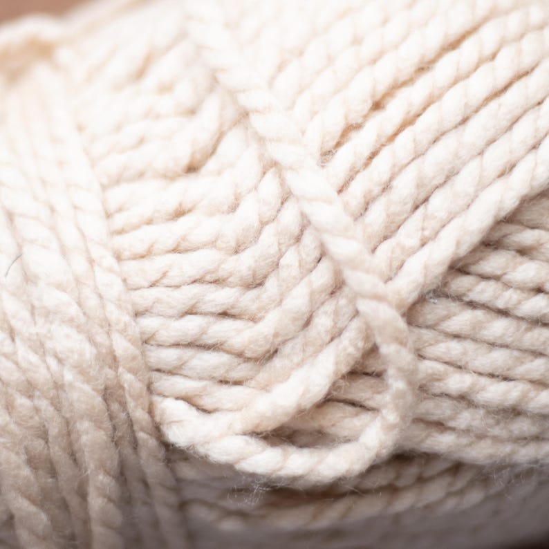 May include: Close-up of a ball of off-white yarn. The yarn is thick and textured, with visible strands tightly wound together. The color is a soft, creamy white, ideal for knitting or crocheting projects.