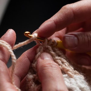 May include: Close-up of crochet work in progress. A gold crochet hook with a yellow handle is used to pull yarn through loops of beige and white yarn, creating a textured fabric. The hands are visible, holding the hook and the work.