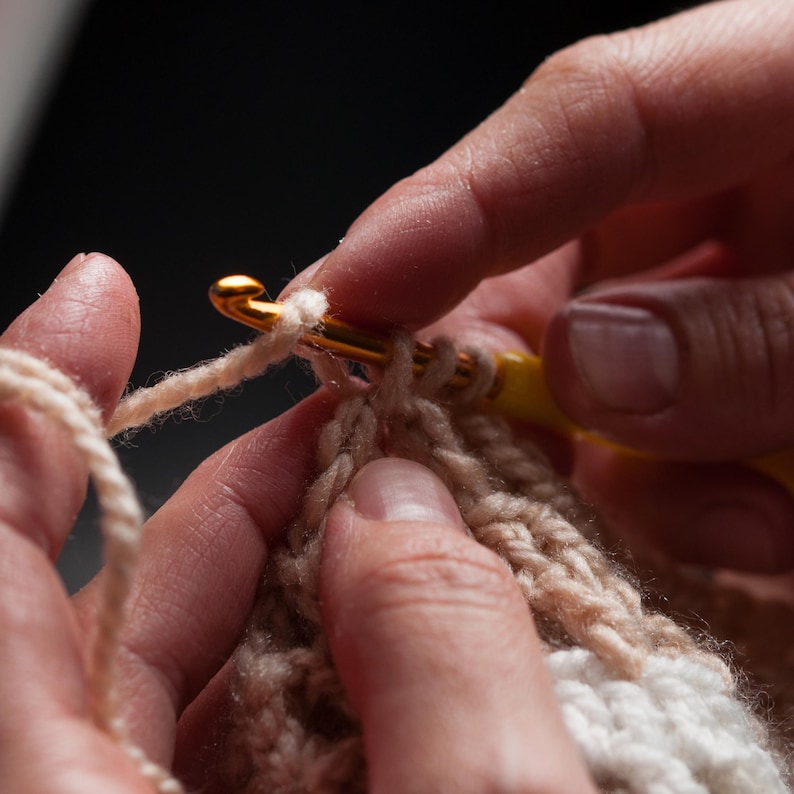 May include: Close-up of crochet work in progress. A gold crochet hook with a yellow handle is used to pull yarn through loops of beige and white yarn, creating a textured fabric. The hands are visible, holding the hook and the work.