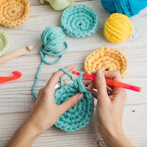 May include: A person's hands crocheting with a teal yarn and an orange crochet hook. Several crocheted circles in yellow, teal, and green are scattered around, along with balls of yarn and crochet hooks, on a light wood surface.