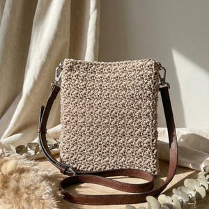 May include: A beige crochet crossbody bag with a brown leather strap. The bag has a rectangular shape and a textured surface. The strap is adjustable and attaches to the bag with silver-colored metal hardware. The bag is displayed on a wooden surface.