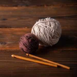 May include: Two balls of yarn, one beige and one dark brown, sit on a wooden surface. Two wooden crochet hooks are positioned diagonally across the bottom of the frame. The yarn balls are different sizes, with the beige ball being larger. The wooden surface has a rich, dark brown color.
