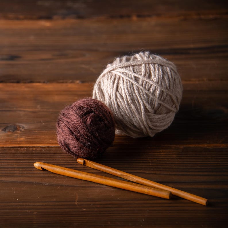 May include: Two balls of yarn, one beige and one dark brown, sit on a wooden surface. Two wooden crochet hooks are positioned diagonally across the bottom of the frame. The yarn balls are different sizes, with the beige ball being larger. The wooden surface has a rich, dark brown color.