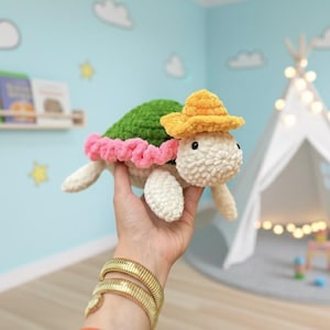 May include: A crocheted turtle toy with a green shell, white head and limbs, and a pink frill. The turtle wears a yellow hat. The toy is held in a hand, with a gold bracelet visible. The background includes a play tent and a light blue wall.