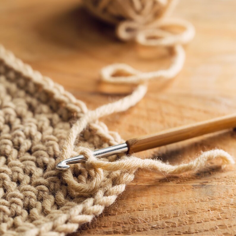 May include: Close-up of crochet work in progress. A wooden crochet hook is inserted into a loop of beige yarn, with a partially completed piece of crocheted fabric visible. A ball of yarn sits in the background on a wooden surface.