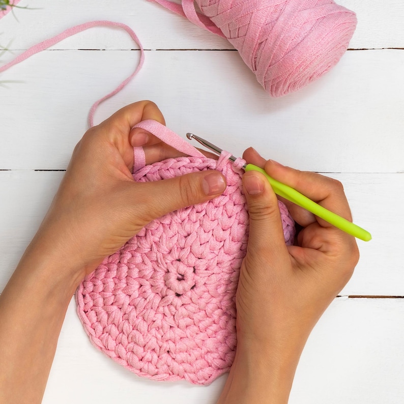 May include: A person's hands crocheting a pink, circular piece of fabric with a green crochet hook. A ball of pink yarn sits in the background on a white wooden surface. The craft project is in progress.