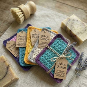 May include: A collection of handmade crochet cloths in various colors, including teal, purple, yellow, and gray, each with a tag that reads "Bebus Design." A wooden brush and bars of soap are also present. The cloths are tied with twine.