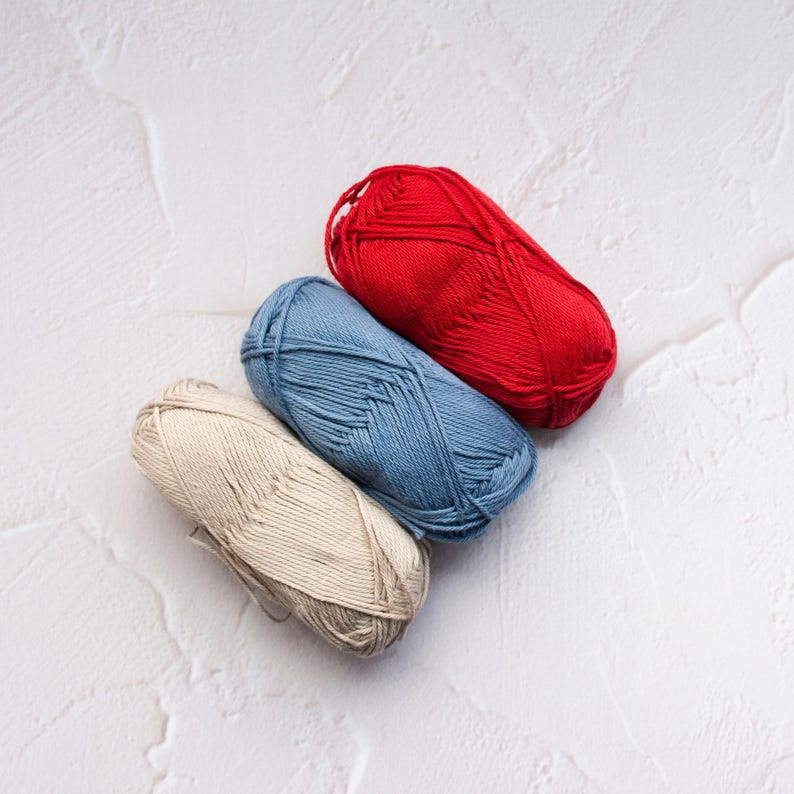 May include: Three skeins of yarn in red, blue, and beige are arranged diagonally on a textured white surface. The yarn is tightly wound, suggesting it's ready for knitting or crocheting projects. The colors offer a versatile palette for various crafts.