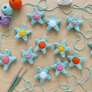 Puede incluir: Estrellas de mar de ganchillo hechas a mano en azul claro, con centros coloridos en rosa, naranja, amarillo y blanco. Las estrellas están dispuestas con hilo y ganchillos sobre una superficie de madera clara, sugiriendo un proyecto artesanal.