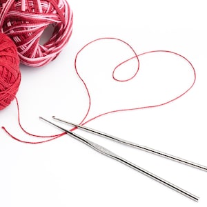 May include: Two silver crochet hooks and red yarn balls on a white background. One ball is solid red, and the other is red and white. A red thread is shaped into a heart, suggesting a crafting theme.