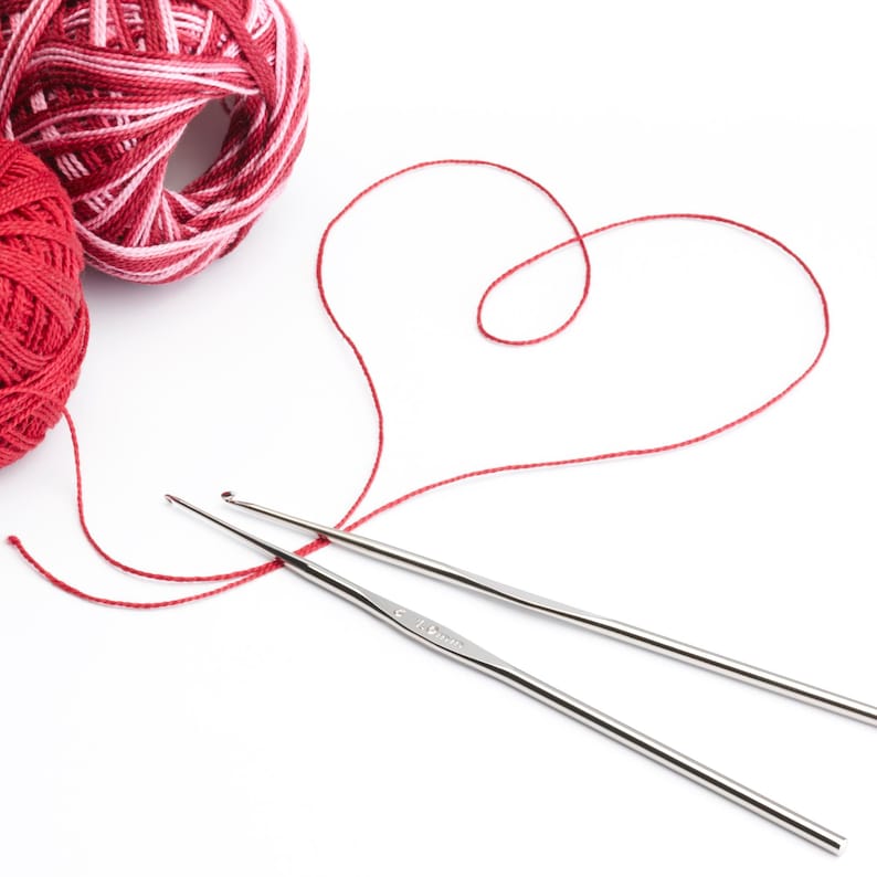 May include: Two silver crochet hooks and red yarn balls on a white background. One ball is solid red, and the other is red and white. A red thread is shaped into a heart, suggesting a crafting theme.