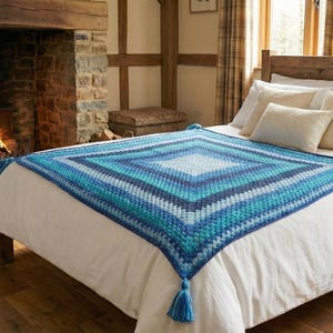 May include: A blue and white crocheted blanket with a diamond pattern is displayed on a bed with white linens. The blanket has tassels at the corners. A fireplace with a burning fire is in the background.