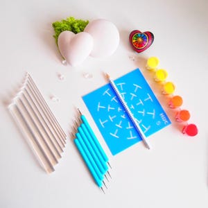 Può includere: Un kit artigianale con vari materiali artistici. Include un oggetto a forma di cuore, un oggetto rotondo, uno stencil blu, una matita, vernice e strumenti per puntinatura. Il kit presenta anche un piccolo oggetto a forma di cuore colorato e oggetti rettangolari trasparenti.