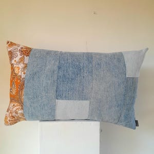 May include: Rectangular cushion with a patchwork design. The cushion features a denim blue centre, light blue patches, and an orange paisley patterned end. The cushion is resting on a white surface.