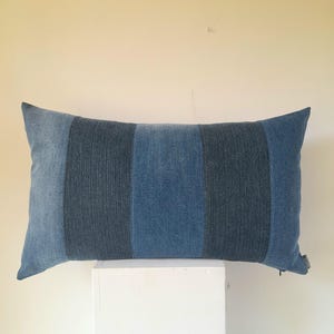 May include: A rectangular decorative pillow with a striped design. The pillow features three vertical stripes in shades of blue, with a zipper closure on one end. The pillow is resting on a white surface against a neutral background.