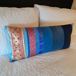 Pode incluir: Uma almofada decorativa retangular com um design de patchwork. A almofada apresenta uma variedade de tecidos de ganga azuis e faixas de tecido coloridas. Uma extremidade tem uma guarnição decorativa com enfeites dourados, vermelhos e verdes. A almofada está sobre uma cama branca.