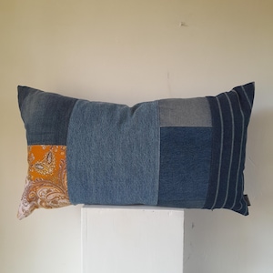 May include: Rectangular patchwork cushion featuring denim and patterned fabric. The cushion's design incorporates various shades of blue denim, with a section of orange paisley fabric. The cushion is approximately 50 cm long.