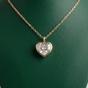 Op de afbeelding: Een gouden ketting met een hartvormige hanger. De hanger heeft een grote, heldere edelsteen gezet in een gouden bezel. De ketting is ook van goud en hangt tegen een groene achtergrond.