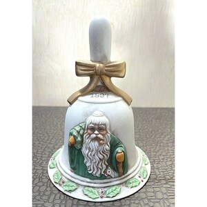 May include: A white ceramic bell with a gold bow and handle. The bell features a depiction of Santa Claus in a green robe, holding a walking stick and a bag of gifts. The base is decorated with green leaves and red berries. The year "1897" is printed on the bell.