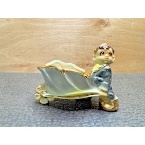 May include: A vintage ceramic planter in the shape of a boy pushing a wheelbarrow. The planter is primarily gray with gold trim and accents. The boy figure has brown hair and is wearing a blue jacket and yellow shirt. The wheelbarrow has a flower-shaped wheel.