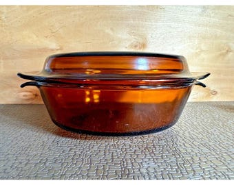 May include: Amber-colored glass casserole dish with a matching lid. The dish has a rounded shape with two handles and a lid that fits snugly on top. The glass has a warm, inviting hue, perfect for serving and storing food.