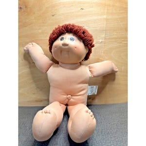 May include: A vintage Cabbage Patch Kid doll with a peach-colored body, arms, and legs. The doll has a round face with rosy cheeks, blue and brown eyes, and a tuft of reddish-brown yarn hair. The doll is sitting with its arms outstretched.