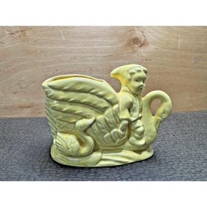 May include: A yellow ceramic planter shaped like a swan carrying a child. The planter has a glossy finish and features detailed wing and feather designs. The child figure is wearing a hooded garment. The swan's neck forms a handle.