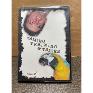 May include: A DVD case titled Taming Training & Tricks Volume 2. The cover features an upside-down photograph of a person's face at the top and a colorful blue and yellow macaw parrot at the bottom, set against a white background with black borders.