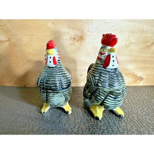 May include: Two ceramic rooster figurines. The roosters have gray and black striped bodies, red combs, and yellow feet. One rooster is slightly larger than the other. The figurines are on a textured surface with a wooden background.