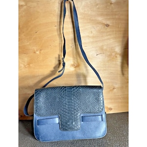 May include: A blue leather crossbody bag with a flap closure. The flap features a snakeskin pattern. The bag has a long, adjustable strap and a rectangular shape. The bag is displayed against a wooden background.