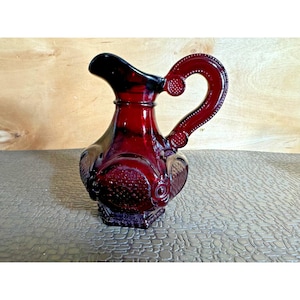 May include: A ruby red glass pitcher with a decorative handle and embossed floral patterns. The pitcher has a hexagonal base and a curved spout. The handle is adorned with small, raised dots. A vintage-style decorative piece.