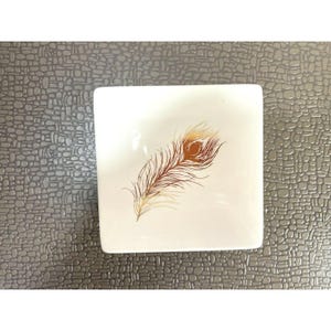 May include: Square white ceramic dish with a detailed peacock feather design in shades of brown, gold, and tan. The feather is centered on the dish, which has a smooth surface.