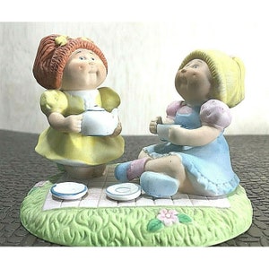 May include: A ceramic figurine depicting two Cabbage Patch Kids having a tea party. One girl wears a yellow dress and holds a teapot, while the other wears a blue dress and holds a teacup. The base is a green picnic blanket with a checkered pattern.