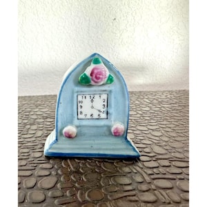 May include: A small, light blue ceramic clock with a square clock face. The clock has a decorative floral design at the top and two pink accents at the bottom. The clock face displays the numbers 1 through 12.