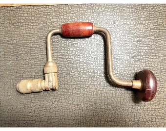 May include: A vintage hand drill with a wooden handle and a metal frame. The drill has a dark brown handle on the top and a matching knob on the end. The metal frame is a silver color. This tool is used for drilling holes in wood.