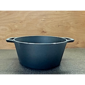 May include: A dark blue cast iron cooking pot with two side handles. The pot has a wide, open design and a textured surface. The pot is sitting on a dark surface with a wooden background.