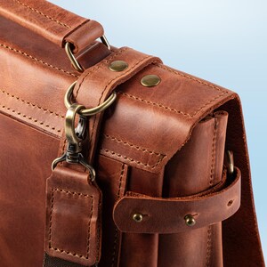 May include: Close-up of a brown leather briefcase with a shoulder strap. The bag features brass-colored hardware, including a clasp, rings, and rivets. The leather has visible stitching and a textured surface, suggesting a high-quality construction.