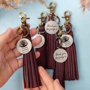 May include: A collection of dark maroon tassel keychains featuring antique brass lobster clasps and circular metal charms. The charms are engraved with a lotus graphic and the text "WOMENS EMPOWER FOUNDATION" or the phrase "thank you for joining us."