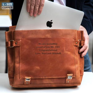 May include: A copper-colored leather messenger bag with a silver laptop inside. The bag has a flap closure with buckles and a front pocket. Text on the bag reads: "We are incredibly proud of you Mr. CEO Congratulations! Love, Burt and Mitchell."