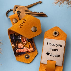 May include: A tan leather keychain with a photo insert and a metal tag. The photo insert shows a man holding a baby. The metal tag says "I love you Pops Austin" with a heart symbol.