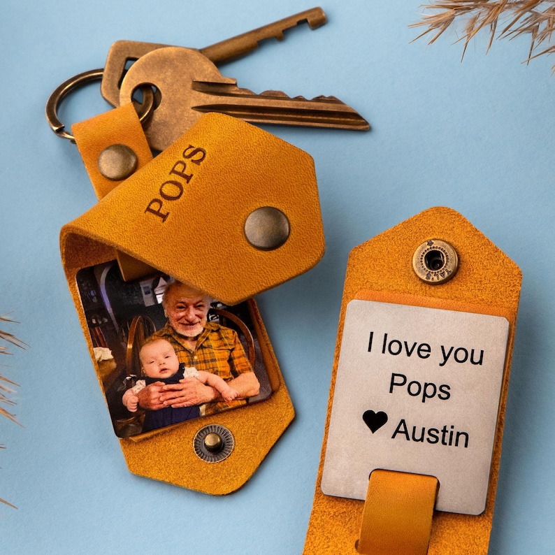 May include: A tan leather keychain with a photo insert and a metal tag. The photo insert shows a man holding a baby. The metal tag says "I love you Pops Austin" with a heart symbol.