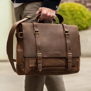 May include: A brown leather satchel with a flap closure, shoulder strap, and top handle. The bag features brass-colored buckles and the text "A.R. Karlin" embossed on the front. The bag is held by a person.